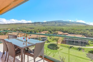A balcony with wicker chairs and a glass table, overlooking a lush landscape with a mountain in the distance. - Maui Resort Rentals: Honua Kai Konea 710 – Top-Floor 1BR with Expansive Mountain Views & Ocean Glimpses (Lahaina)