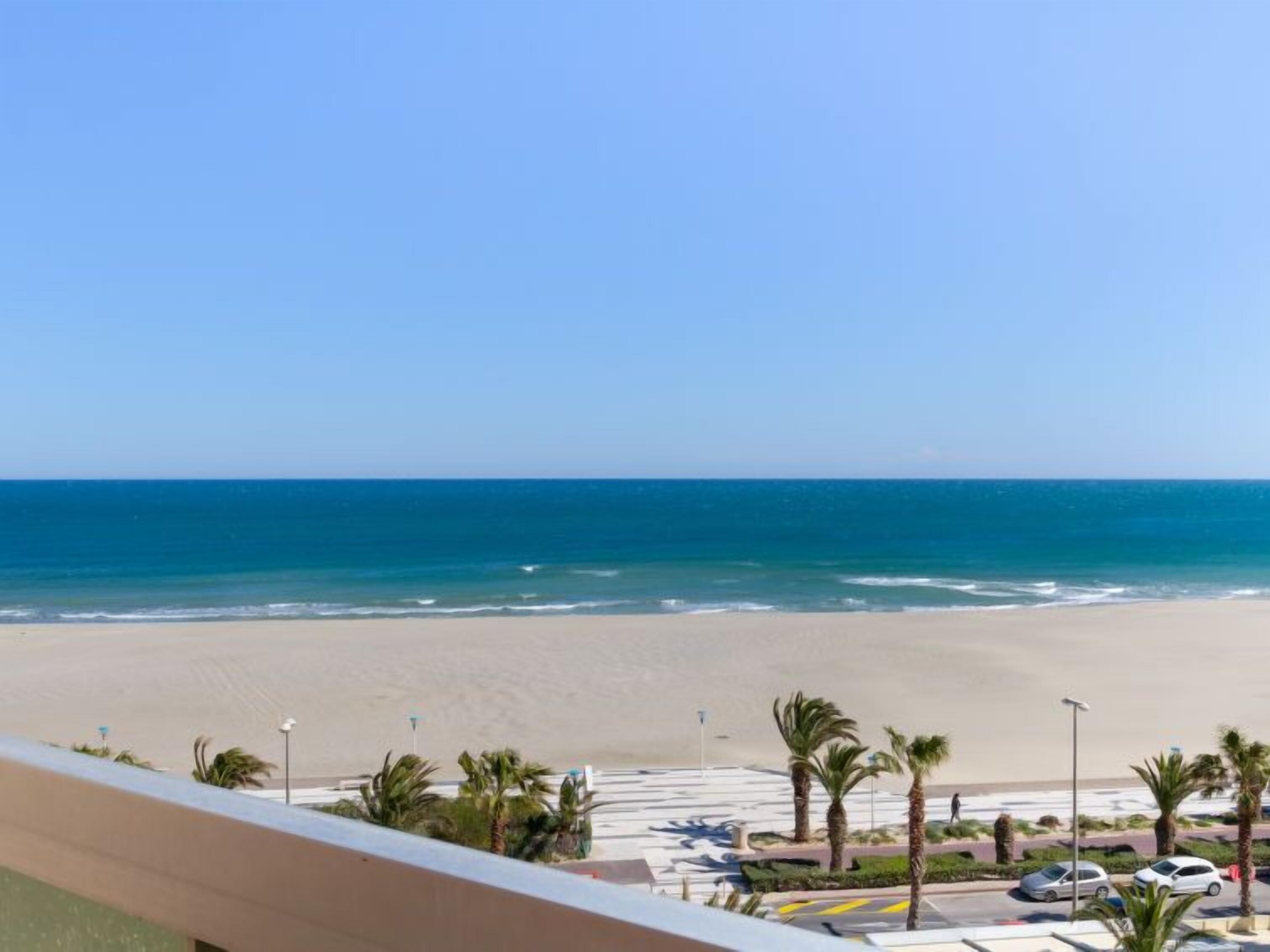 Vista al mar desde un balcón, con un estacionamiento y palmeras al frente.