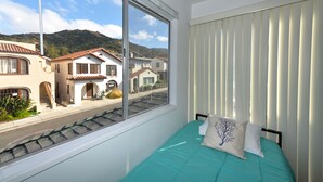 Une chambre avec un lit, une fenêtre donnant sur des maisons et une montagne, et un store.