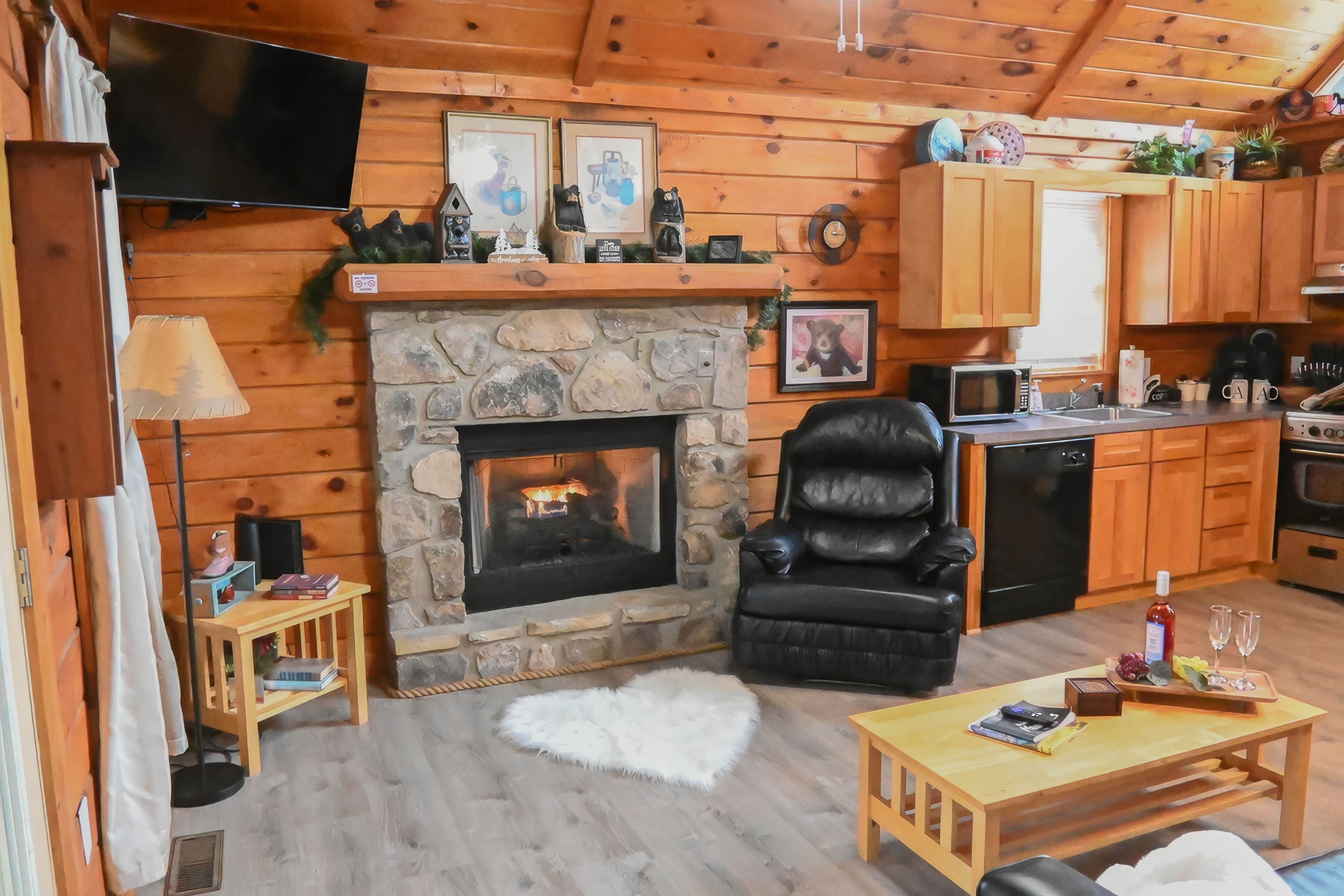 A cozy cabin interior with a stone fireplace, wooden walls, a leather chair, a dining table, and a kitchen area.