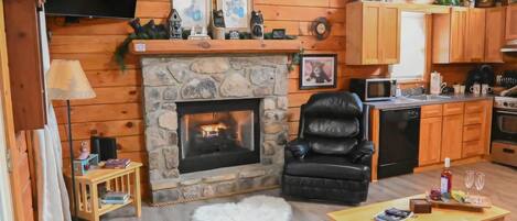 A cozy cabin interior with a stone fireplace, wooden walls, a leather chair, a dining table, and a kitchen area.