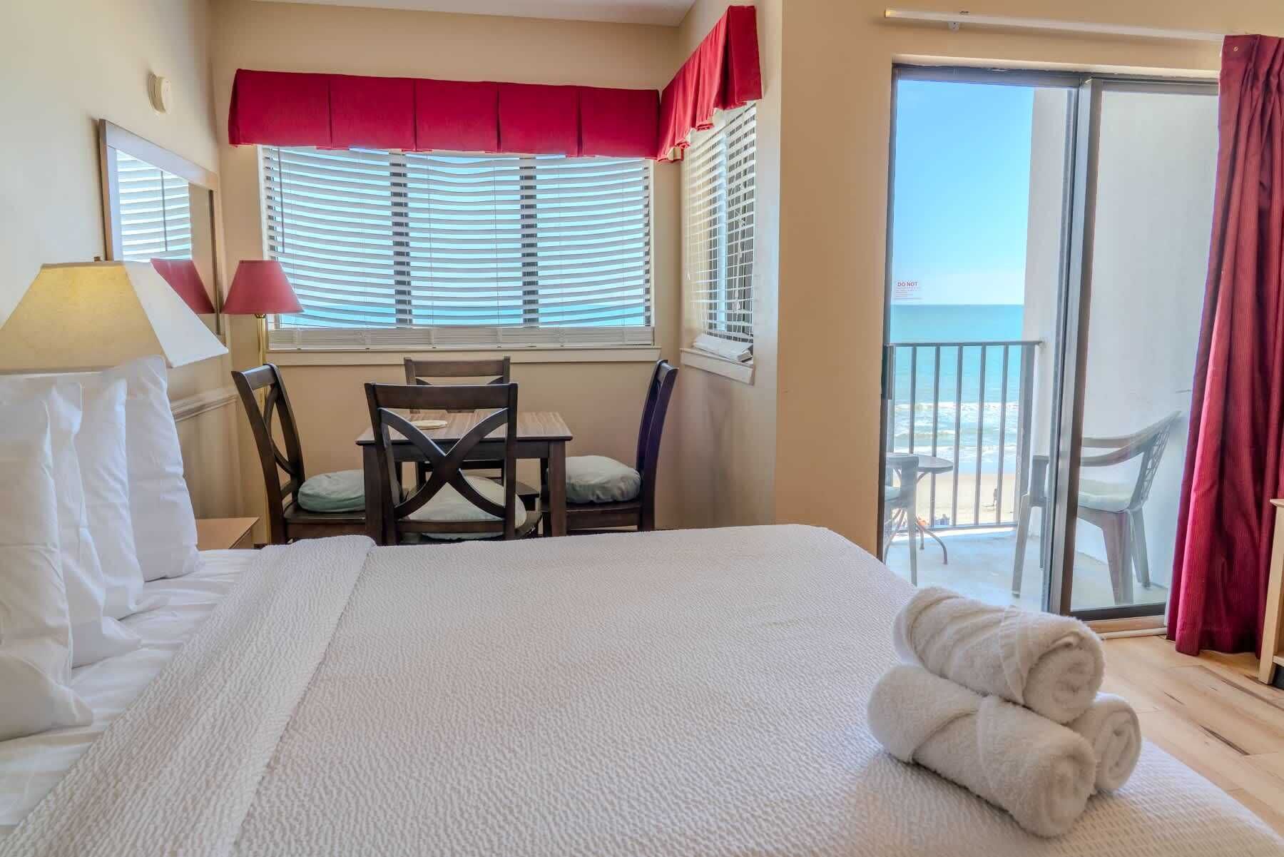 A hotel room with a bed, a table with chairs, a window with blinds, and a view of the beach.