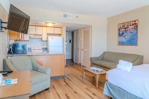 A compact hotel room with a kitchenette, a sofa, a bed, and a painting on the wall.