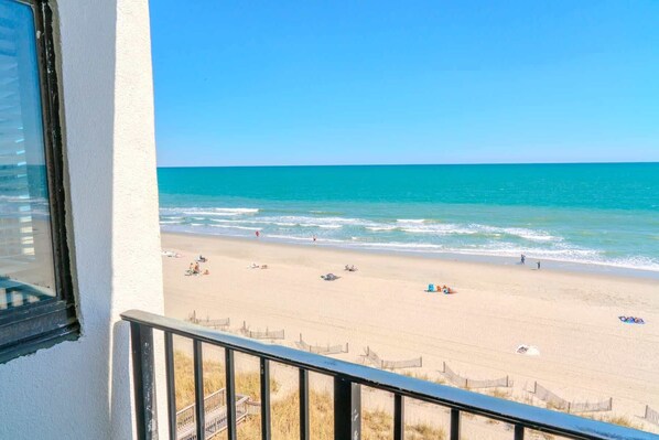 Vista a la playa desde un balcón con barandilla, playa de arena y océano.