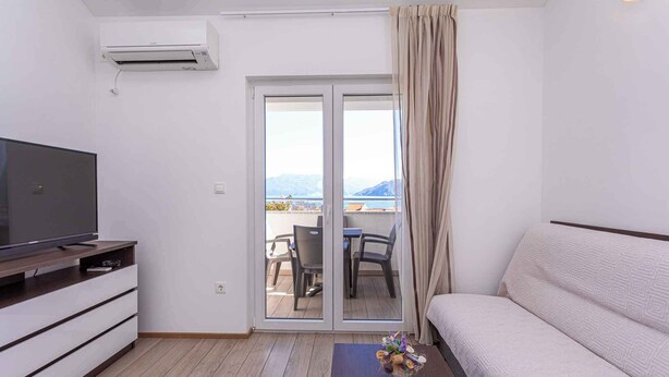 Una sala de estar moderna con una televisión de pantalla plana sobre un mueble de madera, un sofá, una mesita con un florero y un ventanal corredizo que da a un balcón exterior con mesa y sillas.