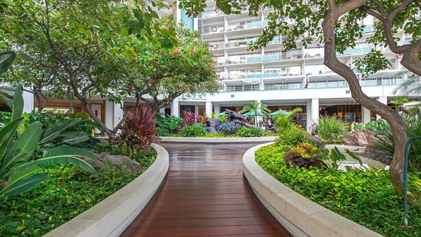 A wooden pathway flanked by lush greenery and a building with balconies in the background.