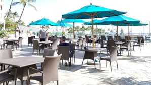 An outdoor café with tables and chairs, blue umbrellas, and palm trees.