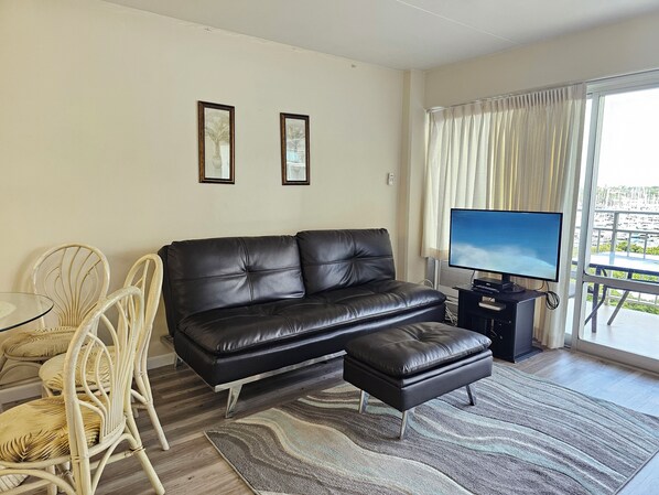 A living room with a black leather sofa, a small ottoman, a glass coffee table, and two white wicker chairs. There is a flat-screen TV on a stand, a large window with curtains, and two framed pictures on the wall.