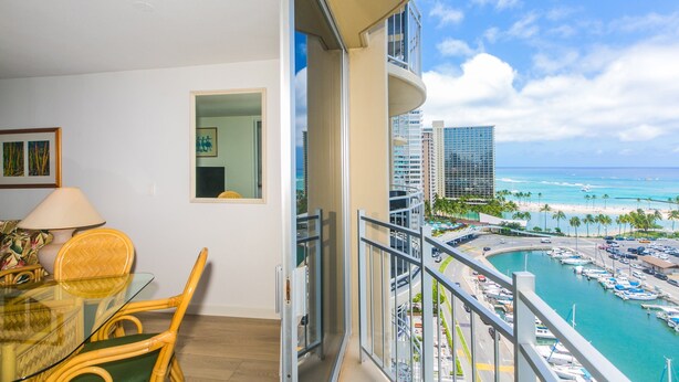 A balcony with a view of a marina and beach, a dining area with wicker furniture, and a modern building in the background. - Breathtaking 180-Degree Ocean Views, Ilikai Marina, Full Kitchen! (Honolulu)