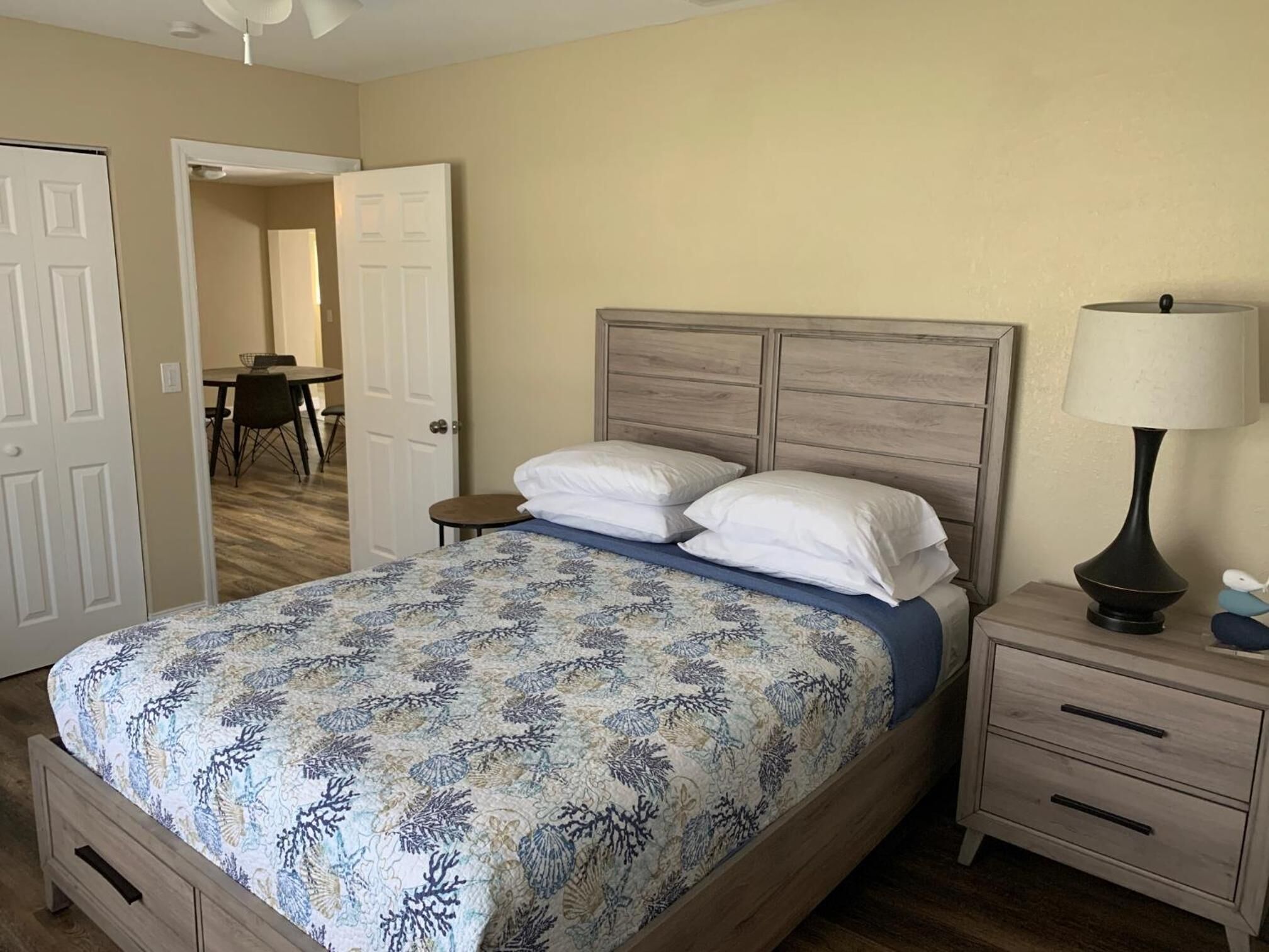 Un dormitorio con cama, mesita de noche, lámpara y puerta que da a un comedor.