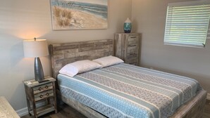 Une chambre à coucher avec un lit en bois, une table de chevet avec une lampe, une commode et un tableau représentant une scène de plage.