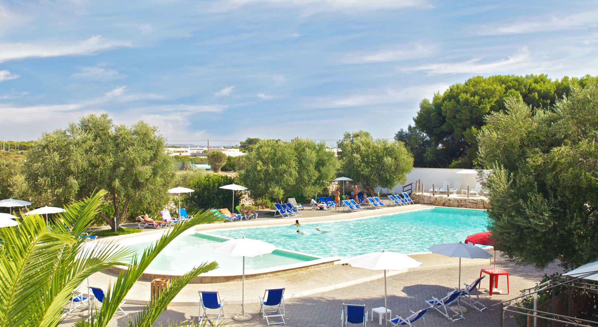 Ein Swimmingpool mit blauen Liegestühlen und Sonnenschirmen, umgeben von Bäumen und blauem Himmel.