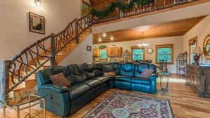 A spacious living area with a large leather sectional sofa, a glass coffee table, a patterned rug, a wooden staircase with a railing, and a view of the kitchen in the background. - THE MOUNTAIN LAUREL |GORGEOUS MOUNTAIN VIEWS (Hiawassee)