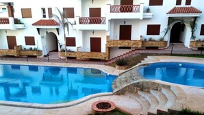 Una piscina con terraza de piedra, un hogar para la leña y un edificio con balcones y tejado de tejas rojas.