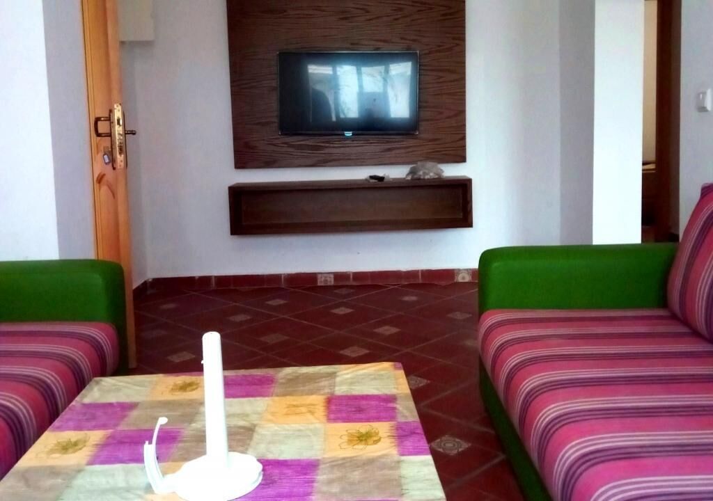 A living room with a television mounted on the wall, a table with a white object, and two green sofas.