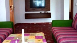 Una sala de estar con un televisor montado en la pared, una mesa con un objeto blanco y dos sofás verdes.