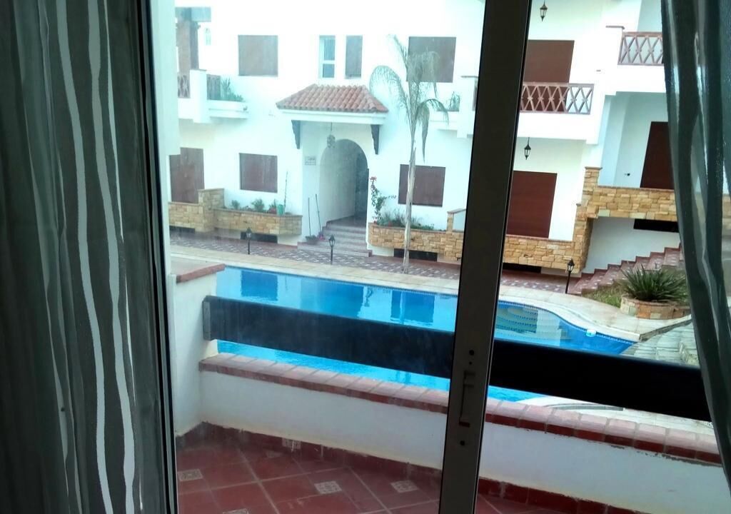 A view of a swimming pool from a balcony with a view of a building with multiple balconies and a staircase leading to an entrance.
