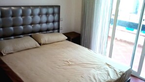 Un dormitorio con cabecera capitonada, una cama grande con ropa de cama color beige, una mesita de noche y vistas a un balcón por medio de un ventanal.