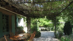 Un patio cubierto con una mesa y sillas de piedra, rodeado de follaje y un sendero de piedra.