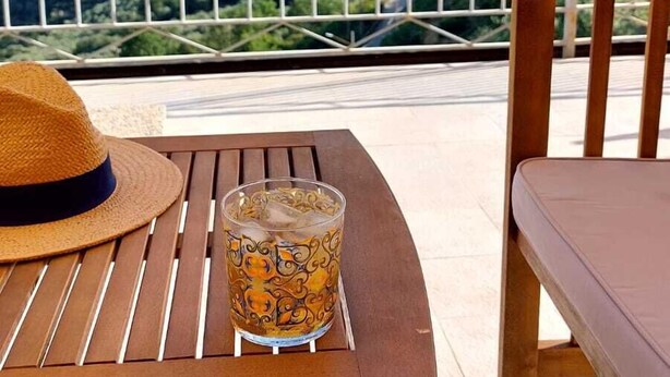 A wooden table with a glass of drink and a straw hat on it, overlooking a scenic view with a metal railing.