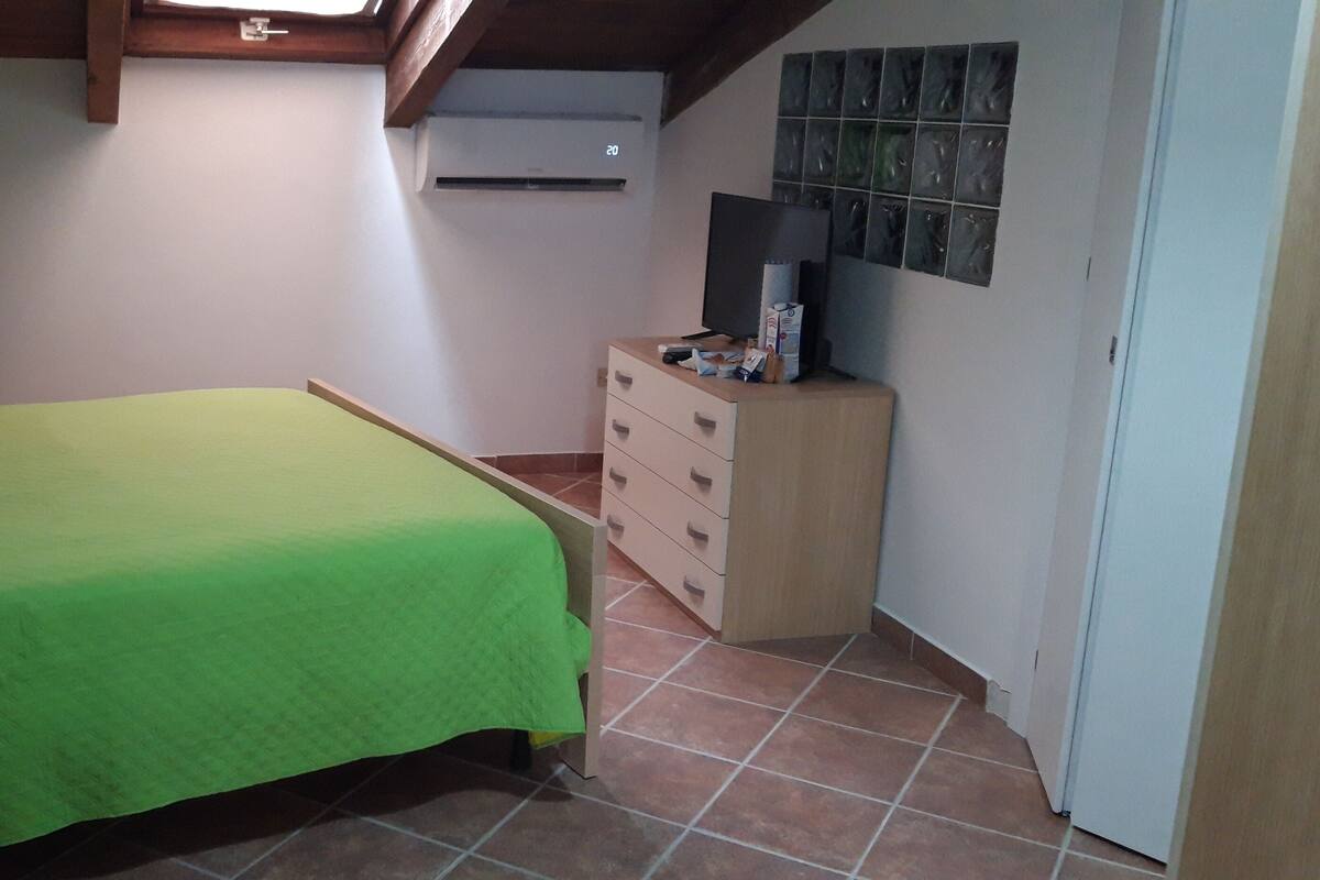Un dormitorio con cubrecama verde, una mesita de noche de madera con lámpara, cómoda y televisor montado en la pared.