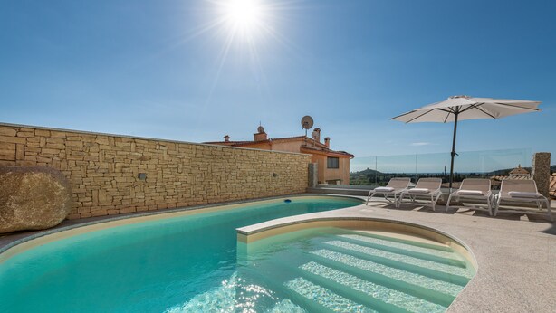 A swimming pool with clear water, a stone wall, and lounge chairs with an umbrella. - Luxurious Holiday Home with Pool, Terrace, Wi-Fi & Air Conditioning; Parking Available, Smaller Pets Allowed (domus de maria)