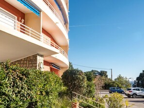 A multi-story building with balconies, a parking area with cars, and surrounding greenery.