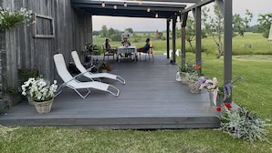 A covered outdoor patio with a wooden deck, white lounge chairs, and tables set for dining. - Newly renovated traditional farmhouse (Ģibuļi)
