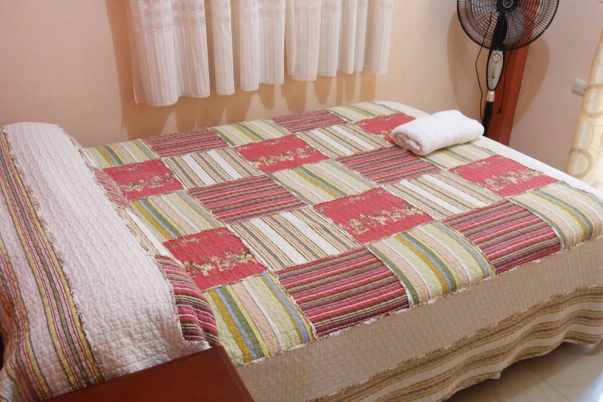 A neatly made bed with a striped quilt, a bedside table, and a standing fan.