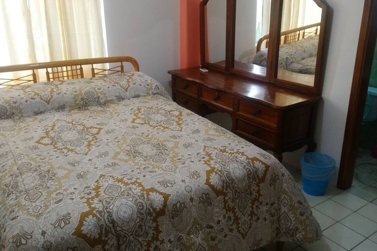A bedroom with a bed, a dresser, a mirror, an air conditioner, and a window with curtains.