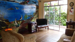 A room with a mural of birds and sea life on the wall, a television, and a large window with a view of greenery. - Hostal Terito (Puerto Baquerizo Moreno)