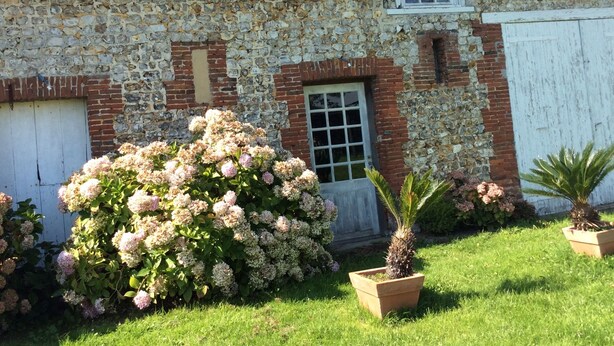 Un edificio de piedra con techo inclinado, una ventana con persiana y una puerta. Un arbusto grande con flores rosas se encuentra frente al edificio.
