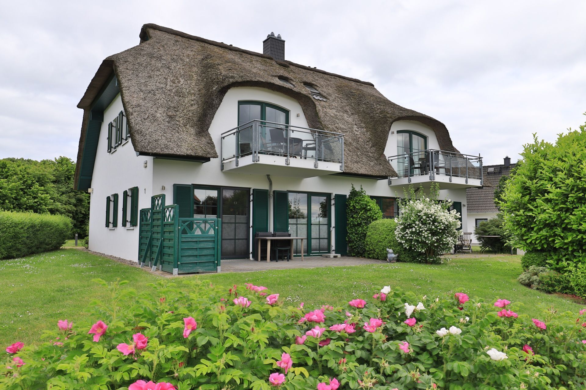 A two-story house with a thatched roof, a balcony, and a garden with pink and white flowers.