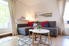 Un salon moderne avec un canapé gris, des coussins rouges et bleus, une table basse ronde en bois et un tableau accroché au mur.