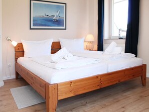 Een tweepersoonsbed met witte lakens, een houten hoofdbord, een ingelijste foto aan de muur, een bedlamp en een raam met gordijnen.