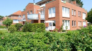 Un immeuble en briques à plusieurs étages, doté de balcons, et entouré de jardins bien entretenus et de plantes en fleurs.