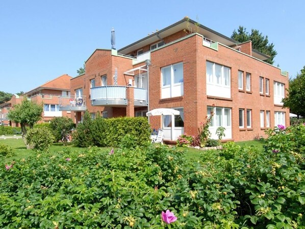 Een meerlaags bakstenen gebouw met balkons, omgeven door verzorgde tuinen en bloeiende planten.
