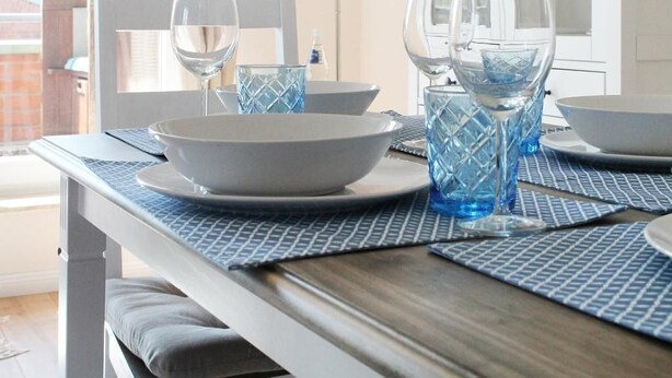 Une table à manger dressée avec de la verrerie bleue et des assiettes blanches, une chaise blanche et une lampe en arrière-plan.