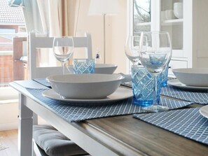 Ein Esstisch ist gedeckt mit blauem Glas und weißen Tellern, ein weißer Stuhl und eine Lampe sind im Hintergrund zu sehen.