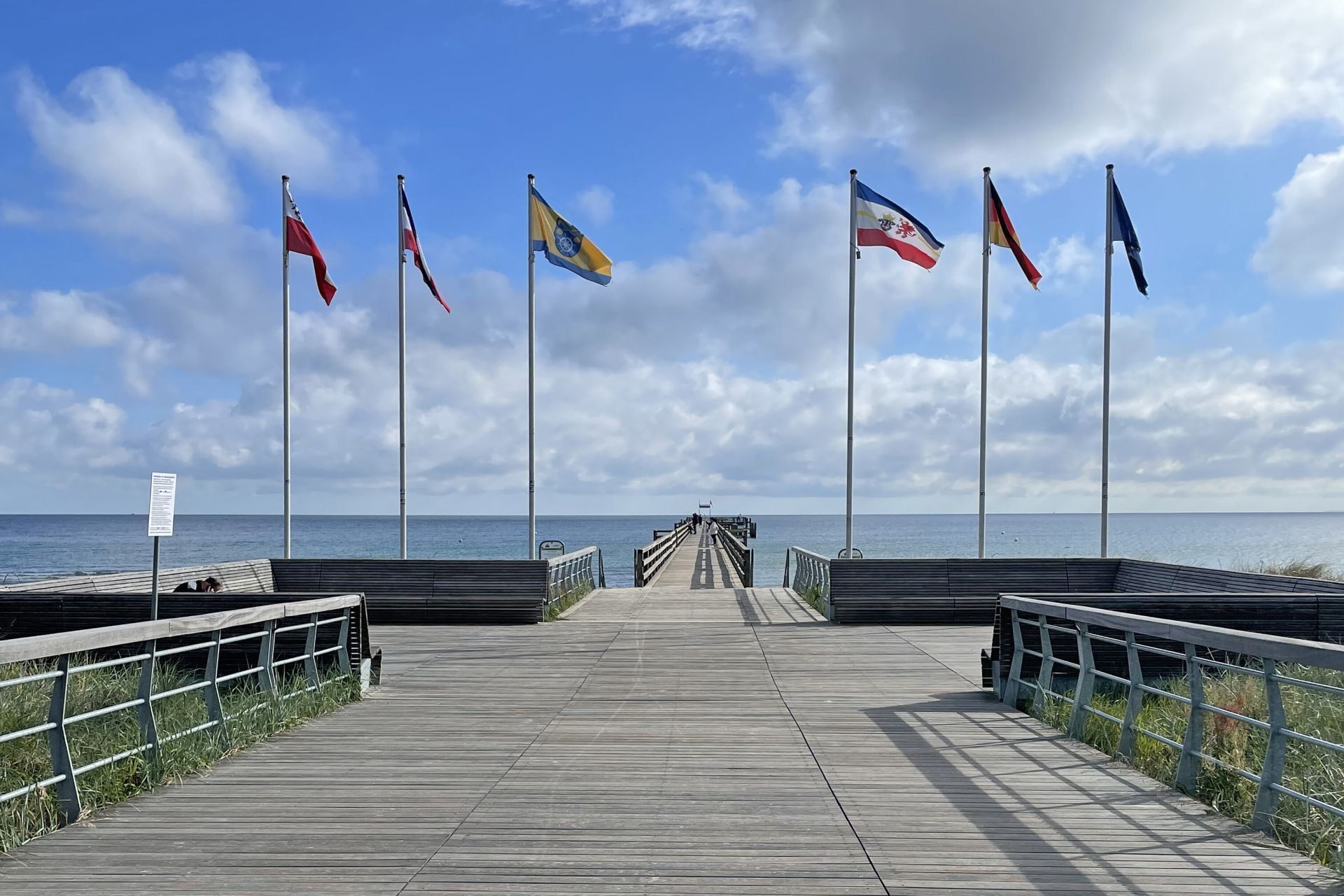 Une jetée en bois qui mène à la mer, ornée de nombreux drapeaux, sous un ciel dégagé, et d’où l’on aperçoit l’océan au loin.