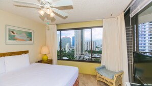 A hotel room with a bed, a chair, a desk with a lamp, a ceiling fan, and a large window with a city view.