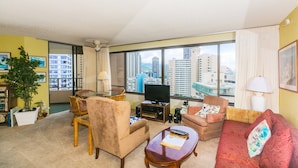 A living room with a television, seating, a plant, and a view of tall buildings.