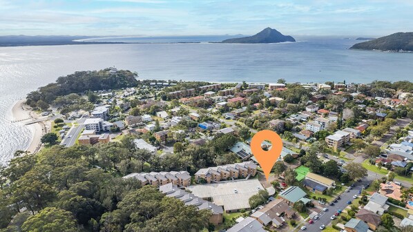 An aerial view of a coastal residential area with a mix of single-family homes and apartment buildings, surrounded by lush greenery and a large body of water.