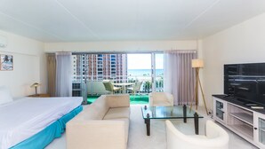 Quarto de hotel com cama, sofá, televisão e varanda com vista para edifícios e para o mar.