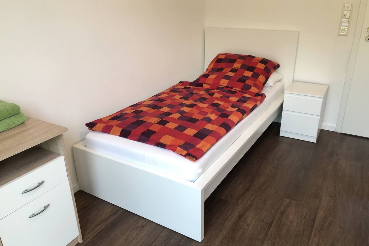 En säng för en person med ett mönstrat överkast, ett sängbord, en liten avlastningsyta och en liten byrå med lådor.