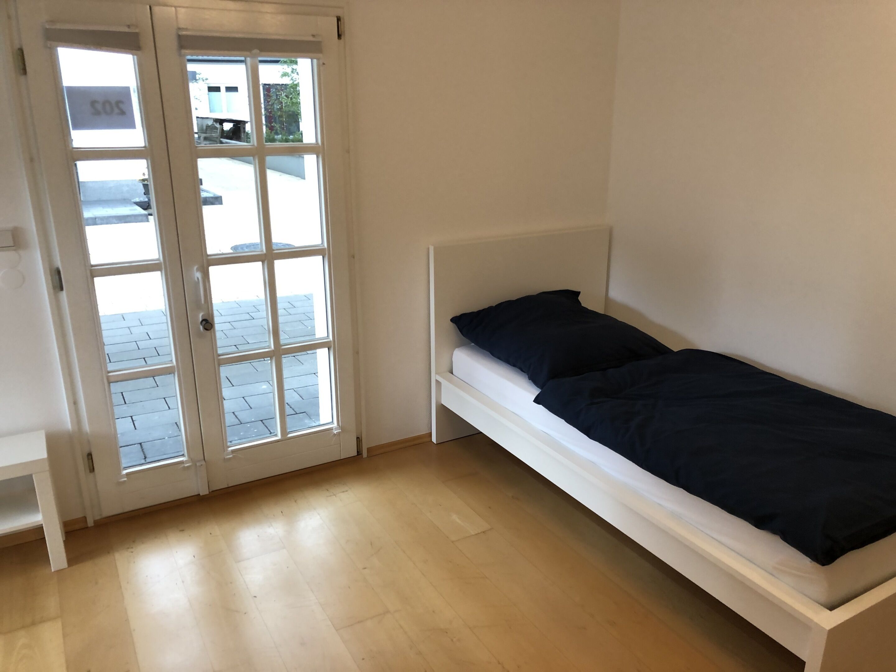 Une petite chambre avec un lit simple, une tête de lit blanche, une fenêtre donnant sur la rue et une porte sur laquelle est affiché un panneau indiquant « SOS ».