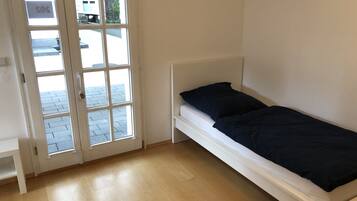Une petite chambre avec un lit simple, une tête de lit blanche, une fenêtre donnant sur la rue et une porte sur laquelle est affiché un panneau indiquant « SOS ».