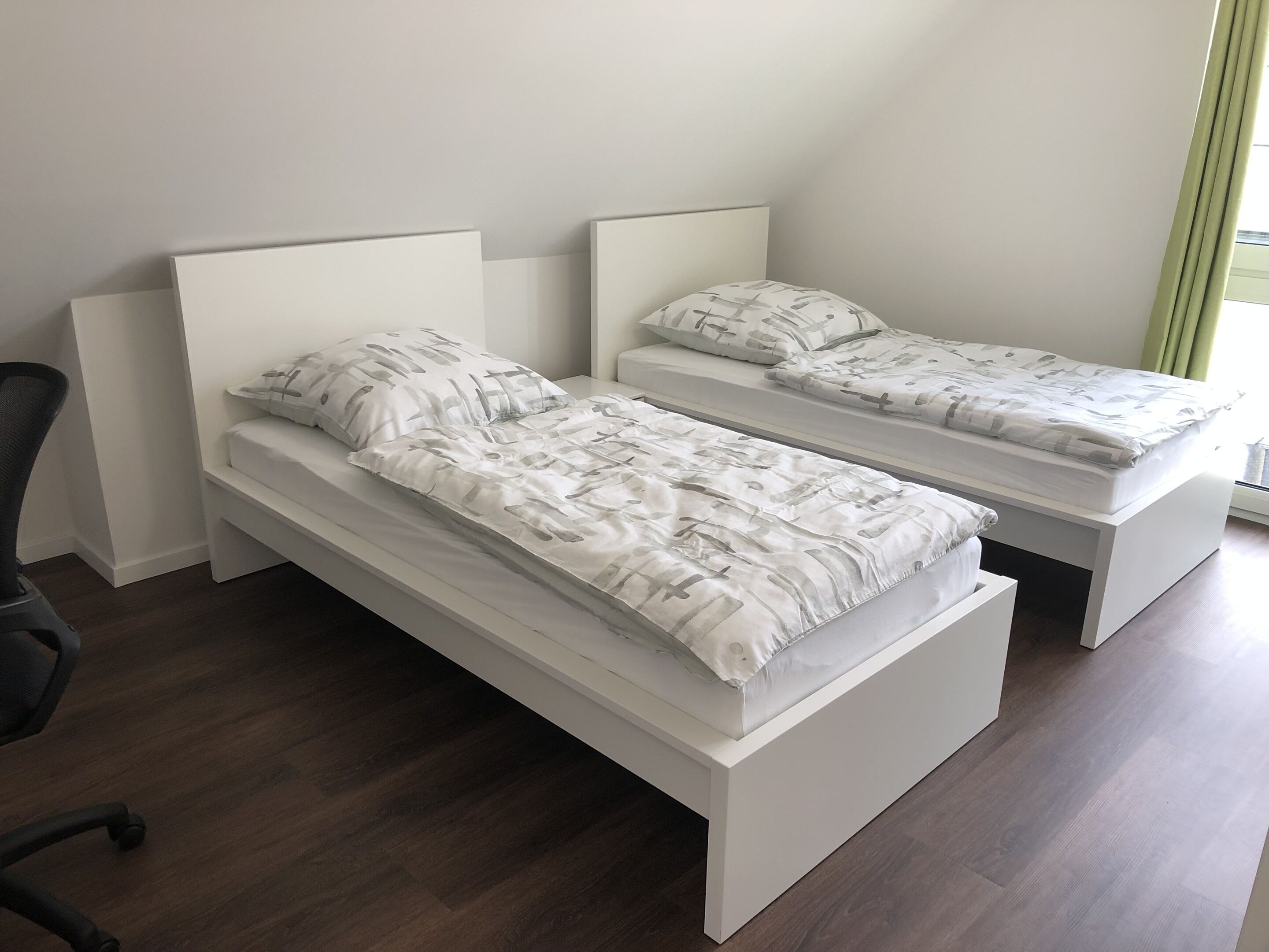 Deux lits simples avec des têtes de lit blanches et une literie à motifs gris dans une chambre dotée d’un plancher en bois et d’une fenêtre avec des rideaux.