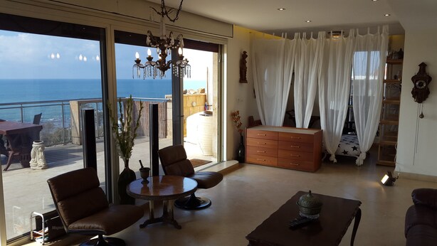 Een ruime woonkamer met een grote schuifpui die uitkomt op een balkon met uitzicht op zee. Er is een kroonluchter, een houten kast en een ronde salontafel.
