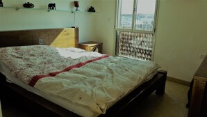 Een slaapkamer met een bed, nachtkastje, raam met uitzicht en een plank met decoratieve objecten.